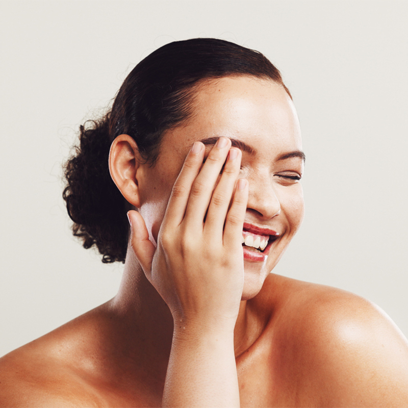 A woman smiling and covering one eye with her hand, against a neutral background.