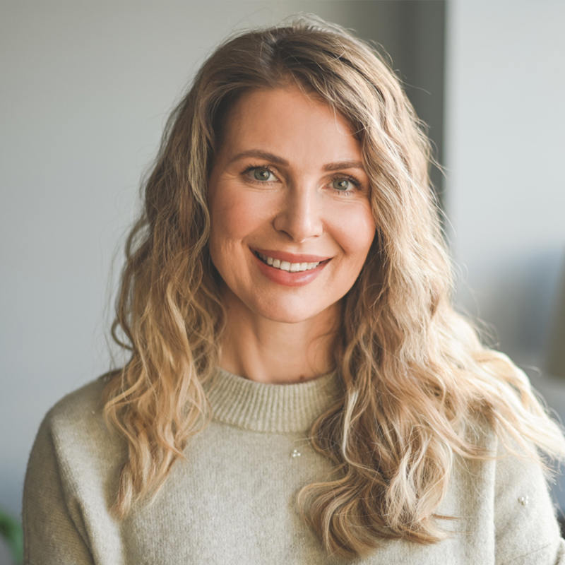 Smiling woman with wavy hair wearing a gray sweater, indoors.