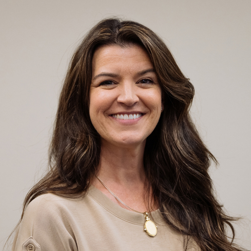 A woman smiling with long brown hair, wearing a beige top and a pendant necklace.