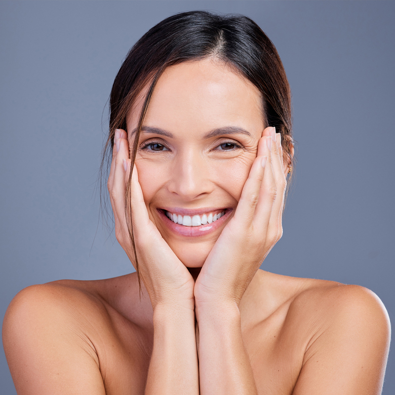 A smiling woman touching her cheeks with both hands on a gray background.