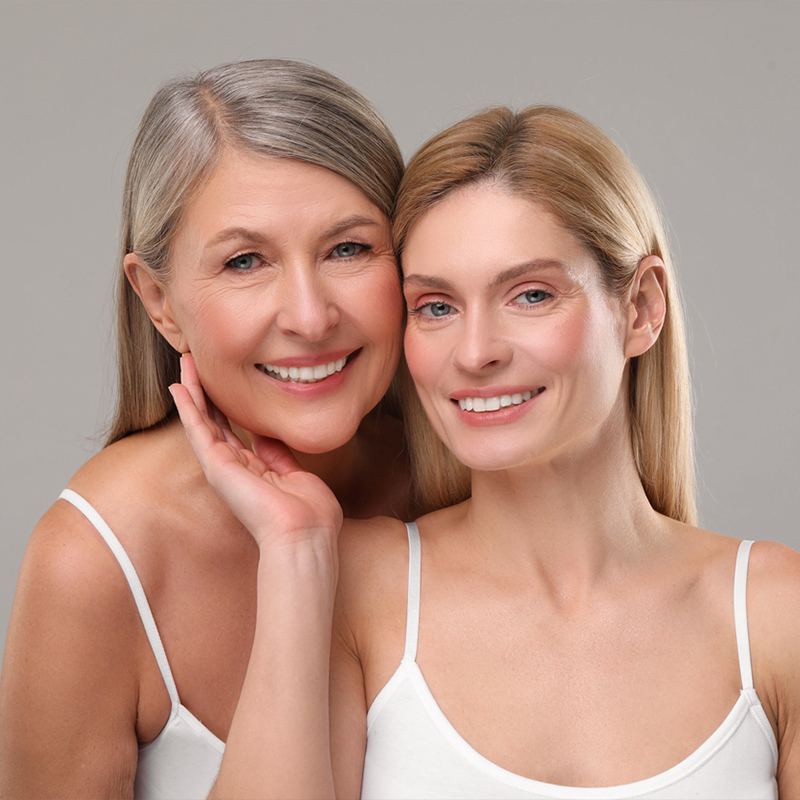 Two women smiling wearing white tops, posing closely in a studio setting.