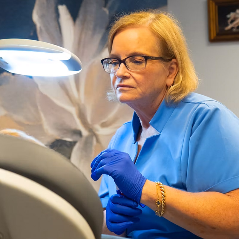 A woman wearing glasses and blue gloves examines something under a lamp in a medical setting.