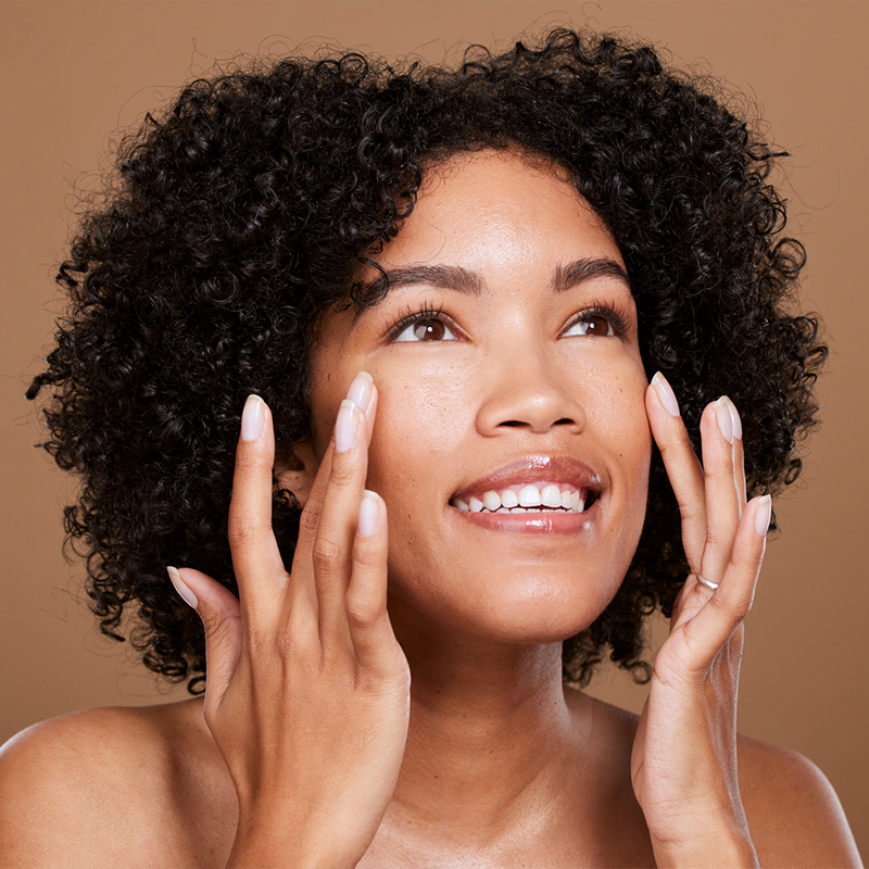 A woman with curly hair smiling and touching her face against a beige background.
