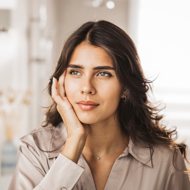 A thoughtful woman with long hair resting her chin on her hand indoors.