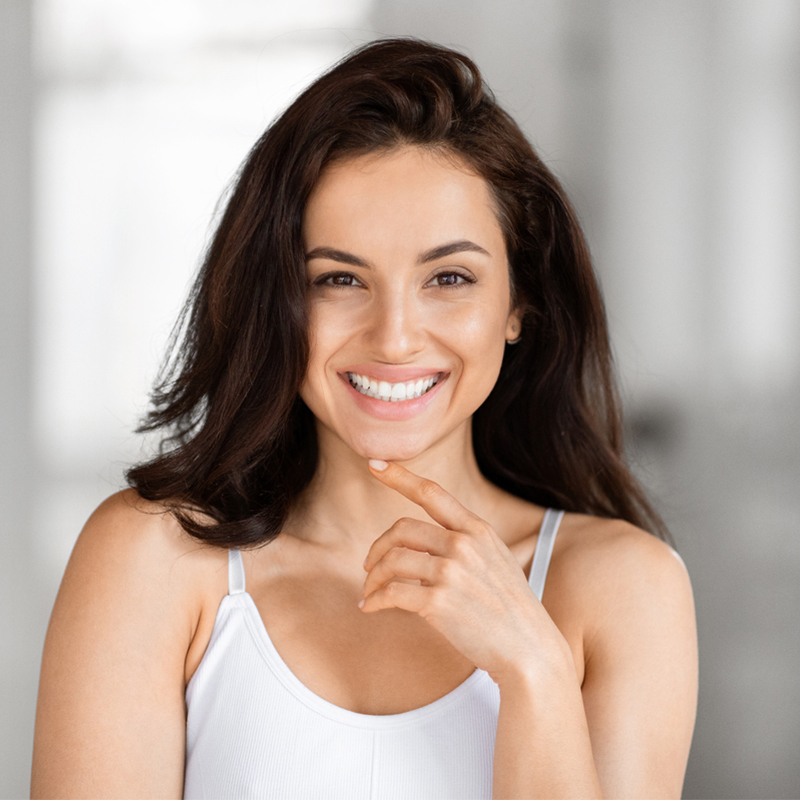 A woman smiling in a white tank top, with a blurred background.