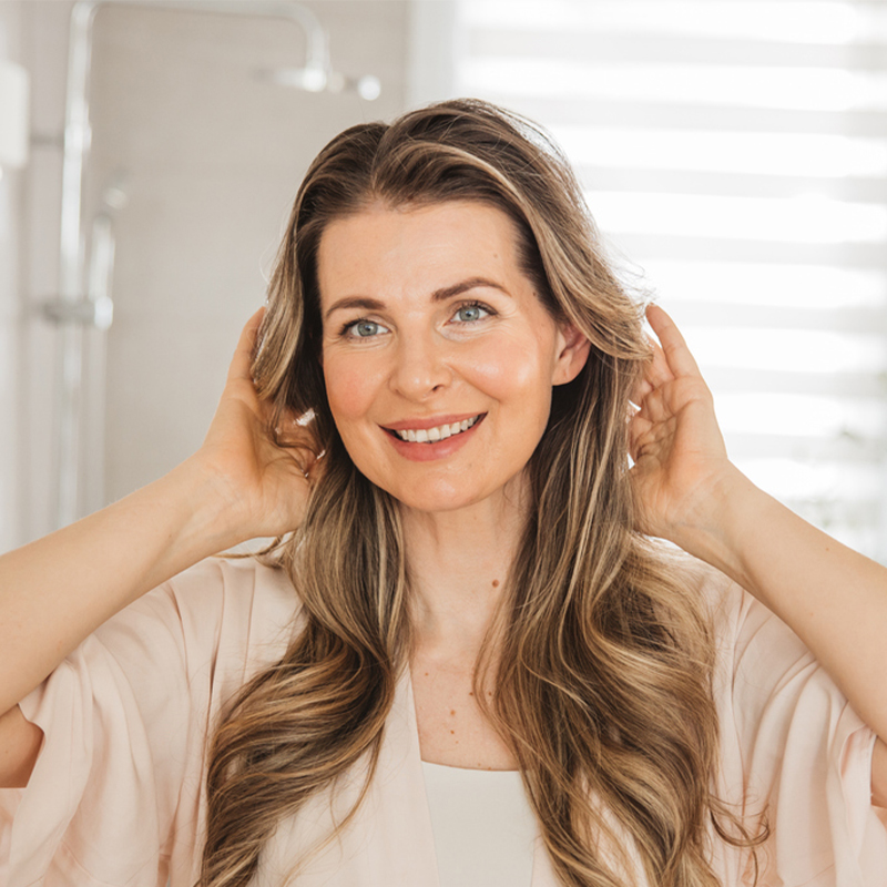 A woman smiling and touching her hair in a bright room.