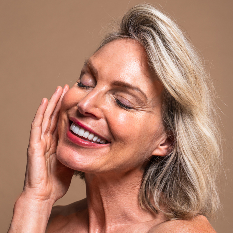 Smiling woman with eyes closed, hand gently touching face against a neutral background.