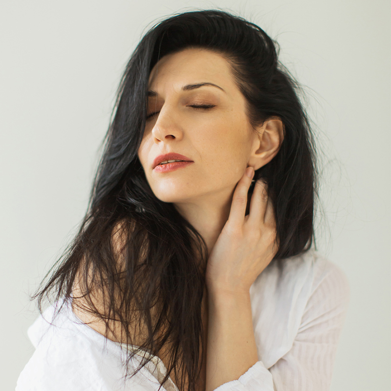 A woman with long dark hair and closed eyes, wearing a white shirt, posing gently with serene expression.