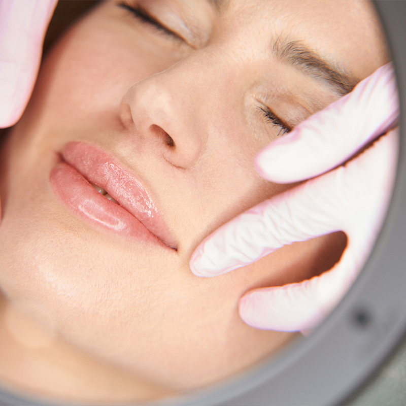 Close-up of a woman's face with closed eyes, touched by gloved hands during a skincare treatment.
