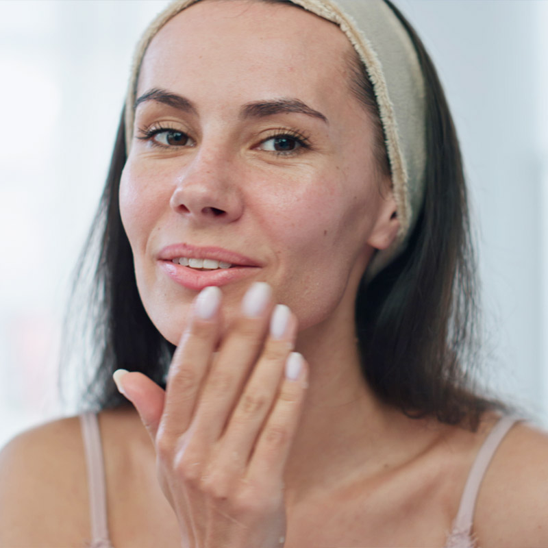 A woman applies skincare product to her face, wearing a headband and smiling slightly.