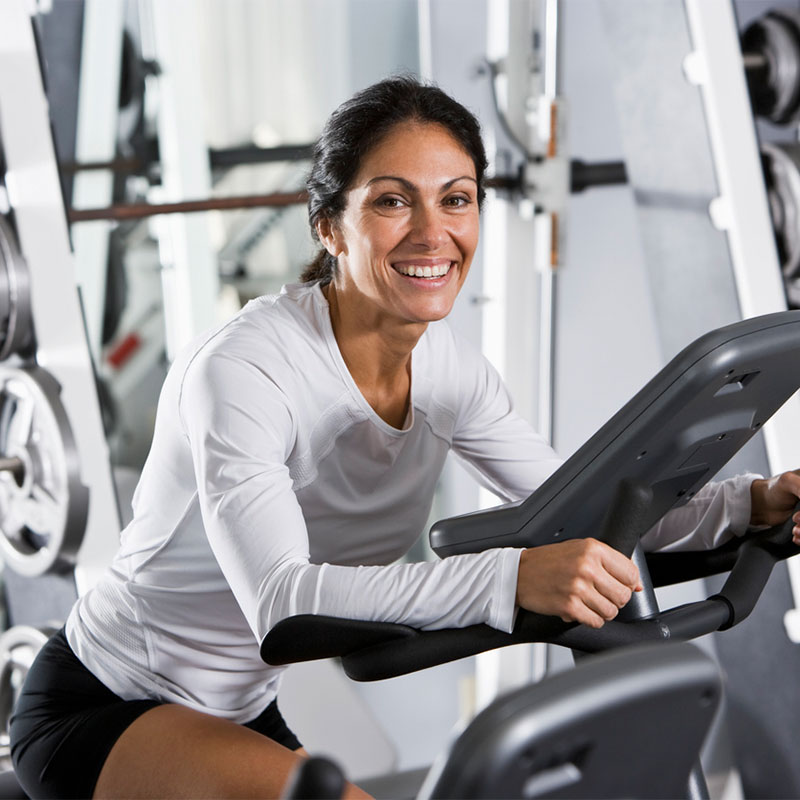 A woman smiling while exercising on a stationary bike at the gym.