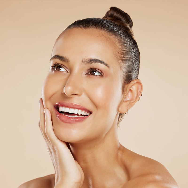 A smiling woman with a bun hairstyle touching her face against a peach background.