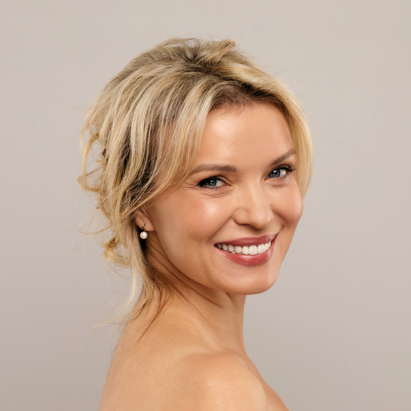 A woman smiling with pearl earrings against a plain background.