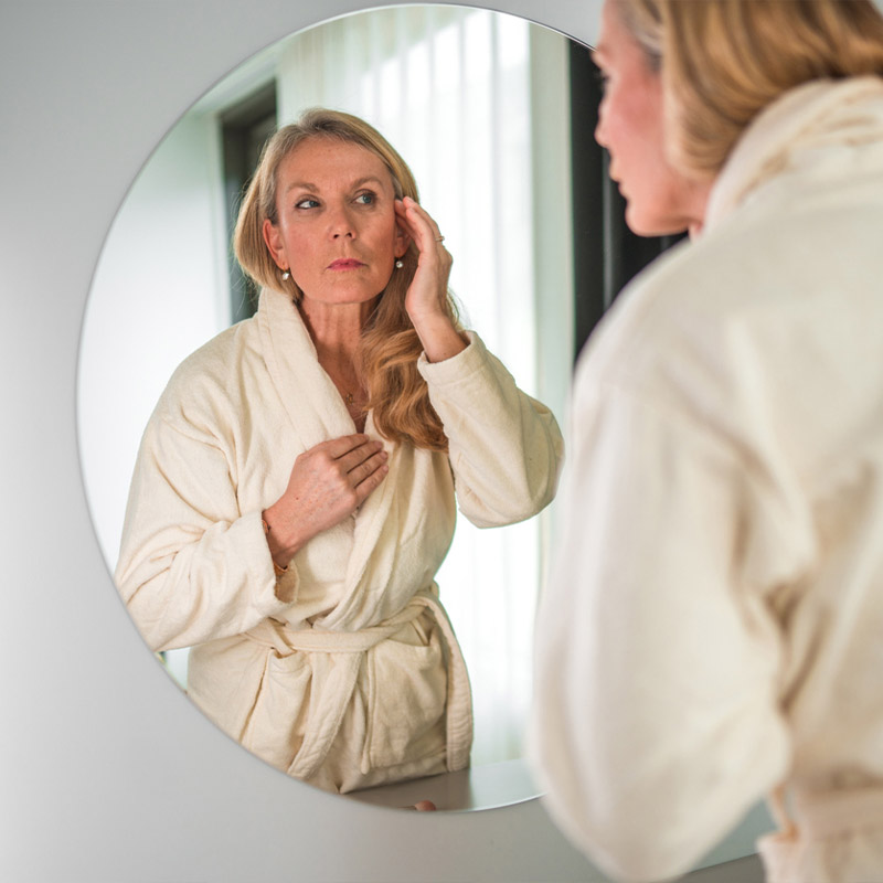 A woman in a bathrobe looks at her reflection in a round mirror.
