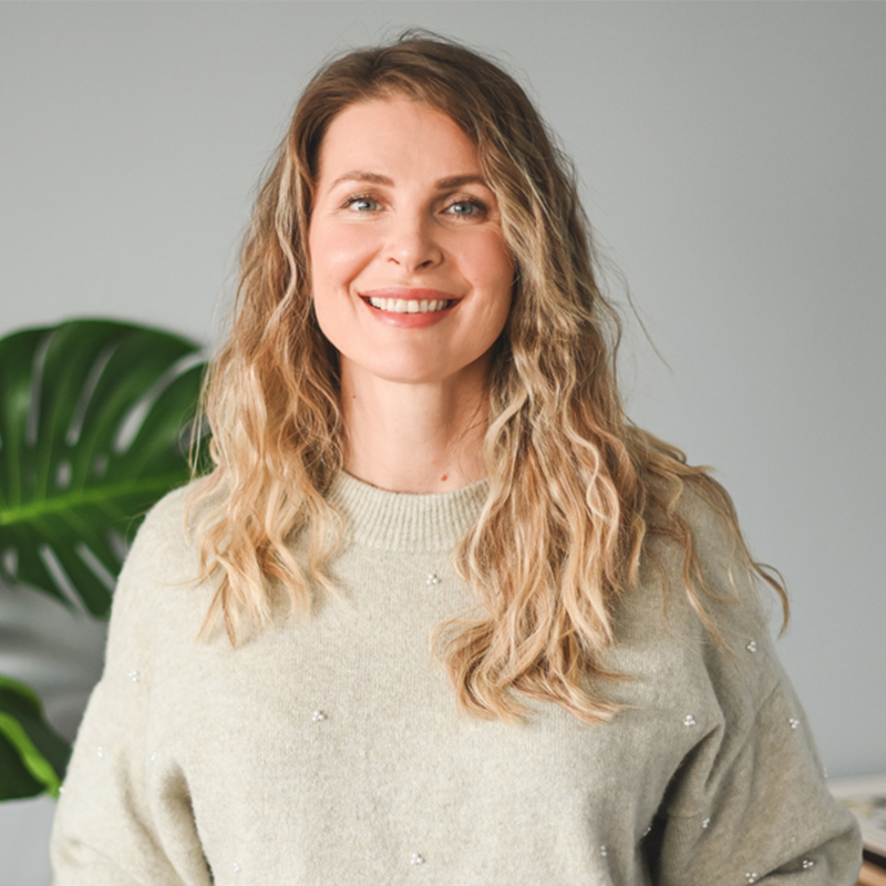 A smiling woman with long wavy blonde hair in a light sweater standing indoors near a plant.