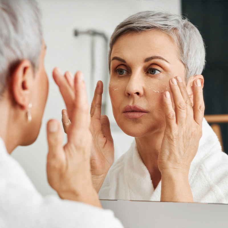 Woman applying cream to face in front of bathroom mirror.